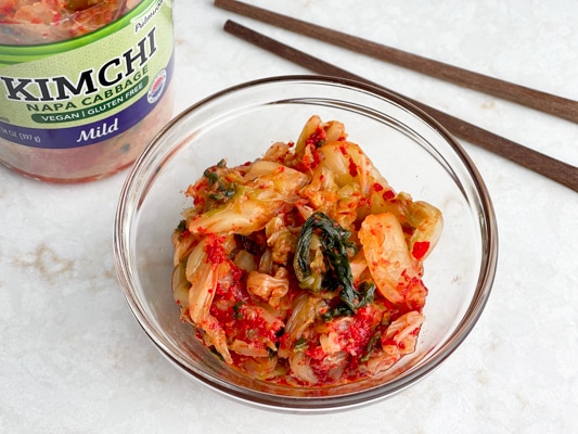 Kimchi in a clear glass bowl with a jar of kimchi and chopsticks on the side.