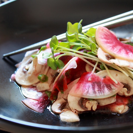 A vibrant mushroom and radish salad in a black bowl with sprouts on top and silver chopsticks on the side