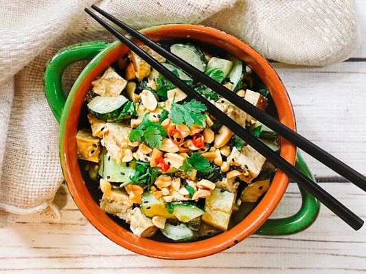 A green bowl filled with a vibrant spicy Asian tofu salad with cucumbers, cilantro, and peanuts, and black chopsticks on top.