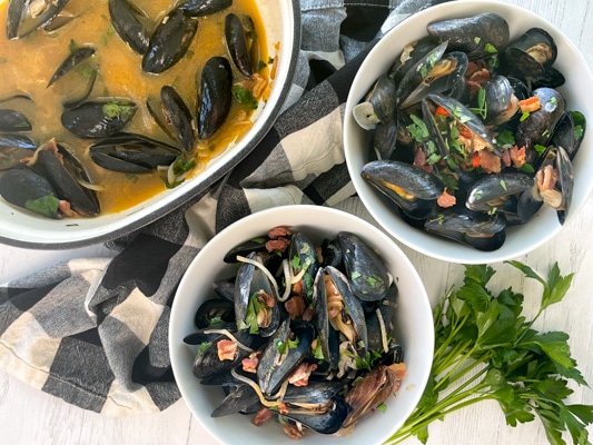 Two white bowls filled with steamed mussels and crusty bread on a white board, with a pot of mussels and green herbs on the side.