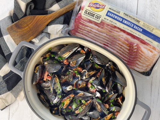 A large pot of steamed mussels tossed with pieces of smoky bacon, sliced shallots, slices of garlic, and fresh green herbs on top of a white board, with a package of Hatfield Smoked Bacon and a wooden spatula on the side.