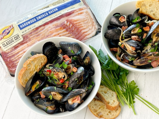 Two white bowls filled with steamed mussels and crusty bread on the side on a white board, with a package of Hatfield Smoked Bacon and herbs and bread on the side.