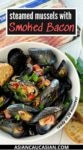 A white bowl filled with steamed mussels on a white board, with a pot of mussels and green herbs on the side.