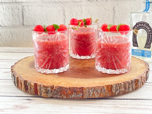 Three crystal glasses filled with boozy watermelon strawberry slushies on top of a rustic wooden tray with a bottle of tequila in the background.