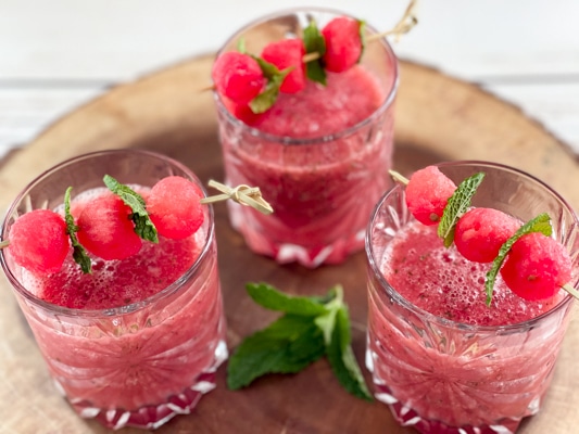 Three crystal glasses filled with boozy watermelon strawberry slushies topped with melon balls, on top of a rustic wooden tray.