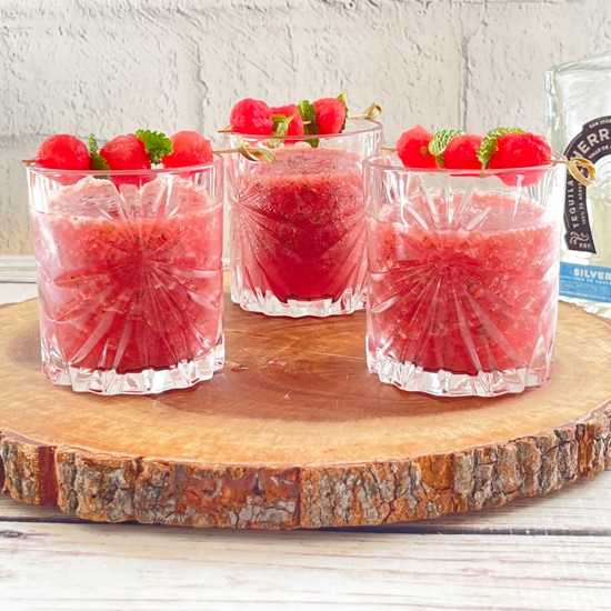 Three crystal glasses filled with watermelon strawberry slushies topped with melon balls, on top of a rustic wooden tray.
