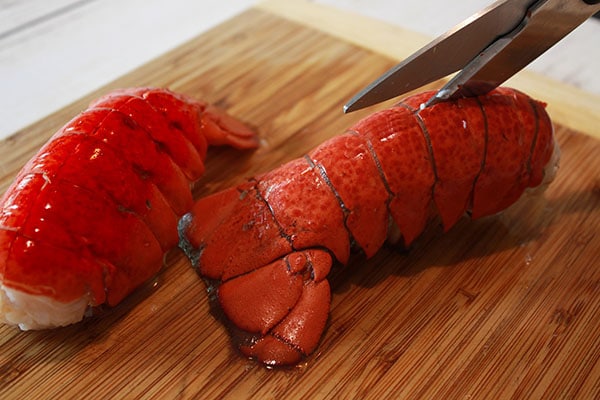 A pair of scissors cutting through the top side of a cooked lobster tail on top of a wooden cutting board.