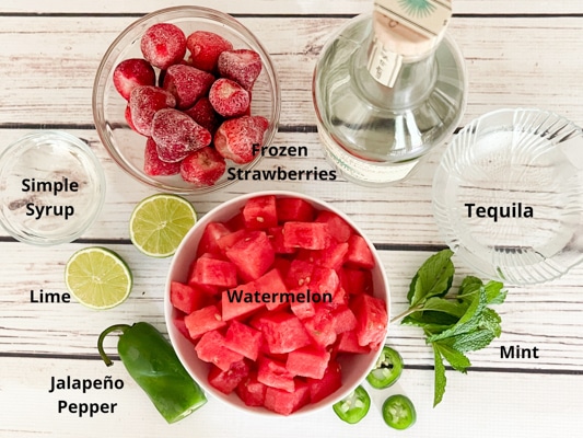 Ingredients on a white board for making boozy watermelon strawberry slushies: frozen strawberries, tequila, watermelon cubes, simply syrup, fresh mint, limes, and a jalapeño pepper.