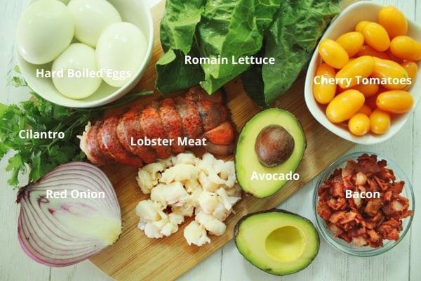Ingredients on a wooden board for lobster cobb salad including, lobster meat, hard boiled eggs, cherry tomatoes, romaine lettuce, red onion, an avocado sliced in half, and pieces of back in a clear bowl.