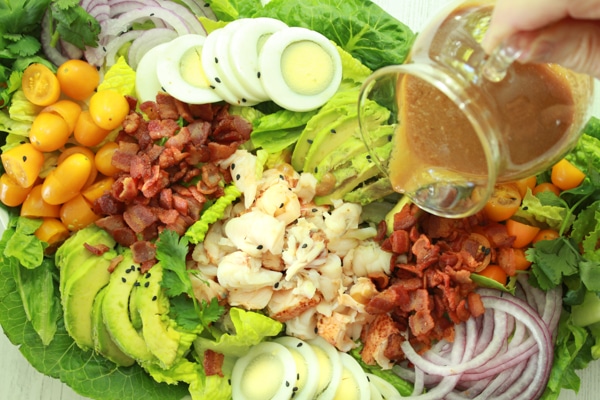 A person pouring miso dressing on top of a vibrant lobster cobb salad with lots of greens.