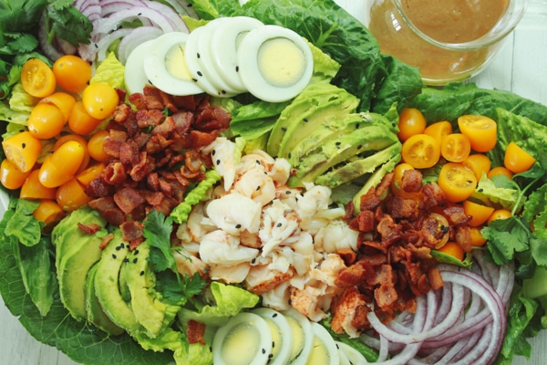 A platter of vibrant lobster cobb salad on top of romaine greens and a clear jar of miso dressing on the side.
