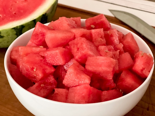 A white bowl filled with fresh watermelon cubes, with a watermelon half in the background.