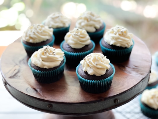 A close-up image of a chocolate miso cupcake with white frosting topped with silver sprinkles, on a wooden board.