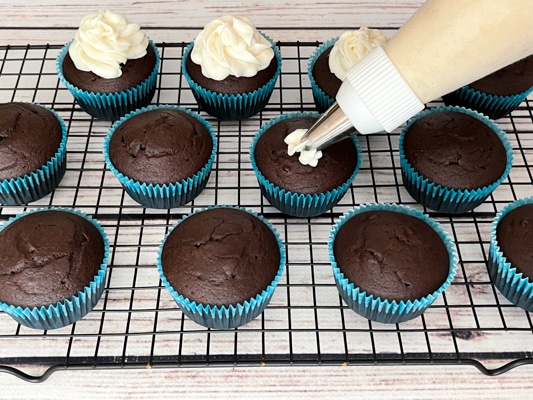 Chocolate miso cupcakes on top of a black wire rack with a piping bag frosting the individual cupcakes.