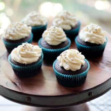 Chocolate miso cupcakes with white frosting topped with silver sprinkles, on a wooden board.