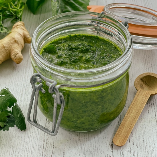 A clear glass jar of cilantro, mint, basil sauce with a small wooden spoon on the side, and bunch of fresh cilantro and basil leaves, and ginger root, all on top of a white wooden board.