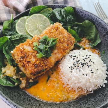 A beautiful piece of golden crusted halibut fillet in a black bowl with a vibrant red curry sauce at the bottom of the bowl, and white steamed rice and spinach on the side.
