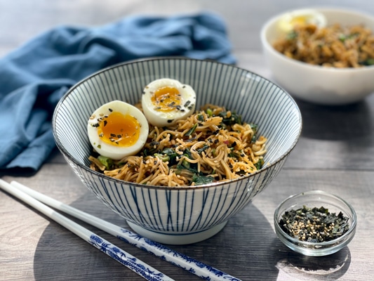 A striped bowl filled with ramen noodles topped with two runny eggs, with chopsticks laying on the side.