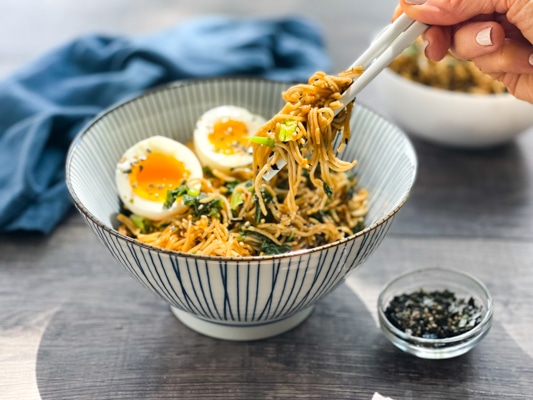 A striped bowl filled with ramen noodles topped with two runny eggs, and a woman's hand with chopsticks picking up some noodles.