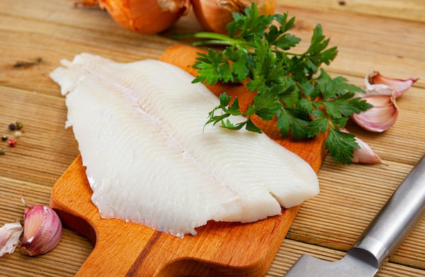 A piece of halibut fillet on a wooden cutting board with a knife, fresh herbs, and garlic cloves on the side.