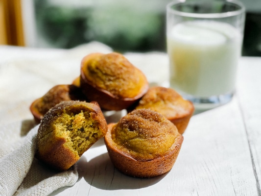 A stack of pumpkin muffins on a white board with a glass of milk in the background and a napkin on the side.