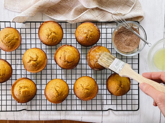 Freshly baked pumpkin muffins on a wire baking rack with a woman brushing the tops with butter.