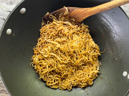 Ramen noodles frying in a large black wok, with a wooden spatula inserted inside.