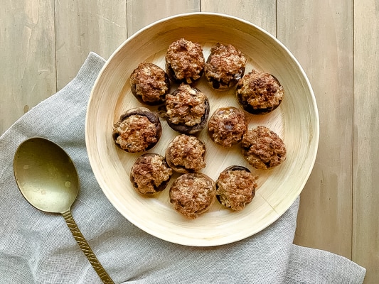 Classic stuffed mushrooms on a bamboo bowl on top of a wooden board with a bronze spoon and linen napkin on the side.