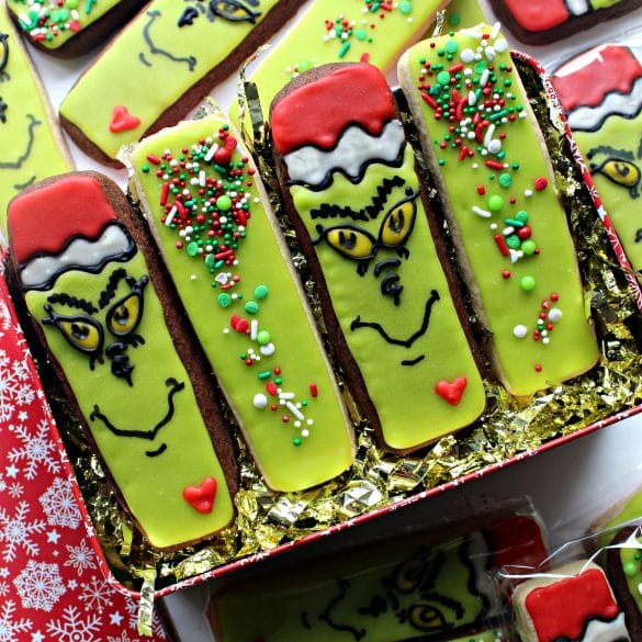 Beautifully decorated Christmas Grinch cookie sticks lined up in a cookie tray on top of shredded gold paper.