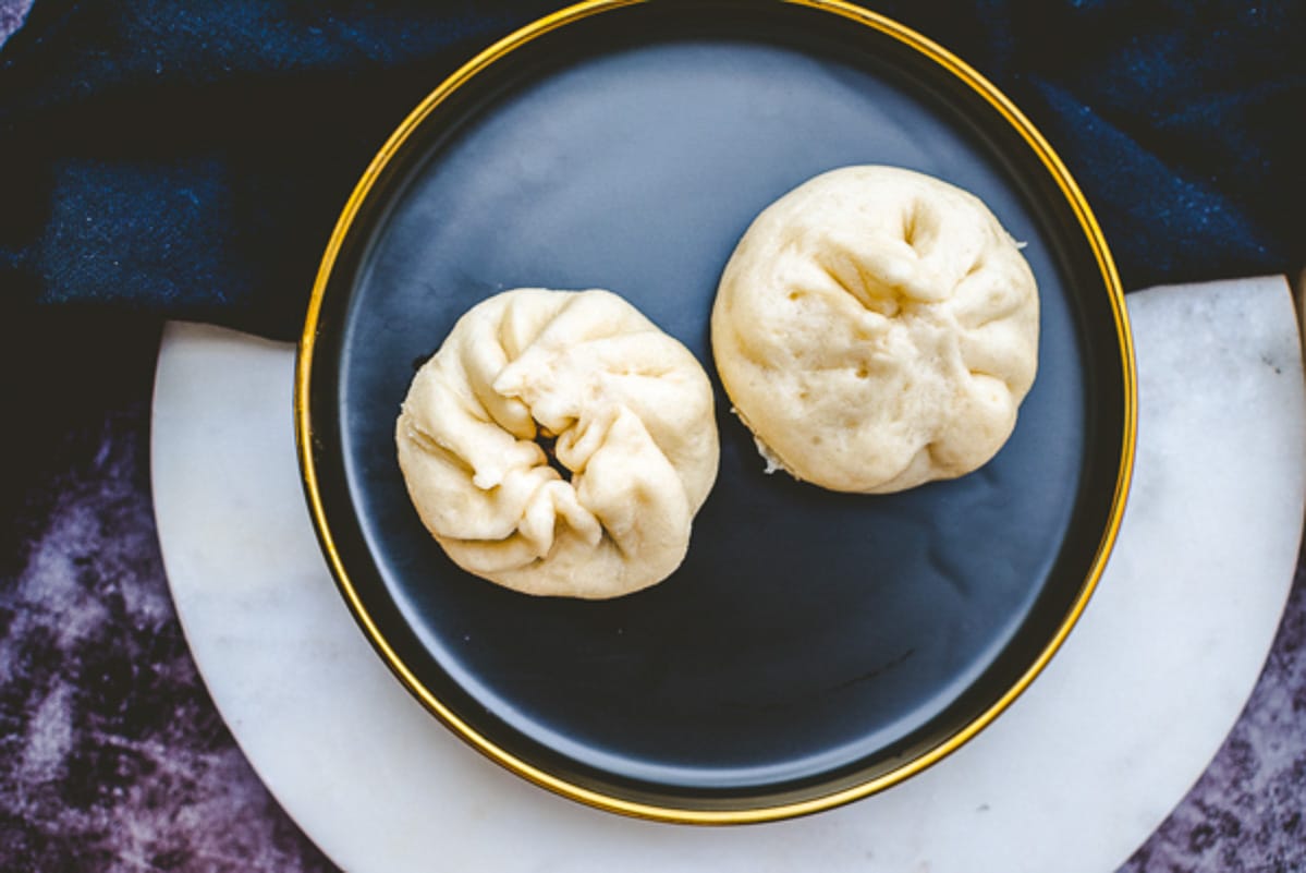 Two Chinese steamed pork dumplings on a dark round plate on top of a round marble serving platter.