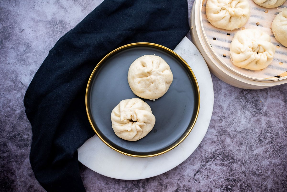 Two Chinese steamed pork buns on a round black plate with a bamboo steamer on the side filled with steamed buns, on top of a gray marbled surface.