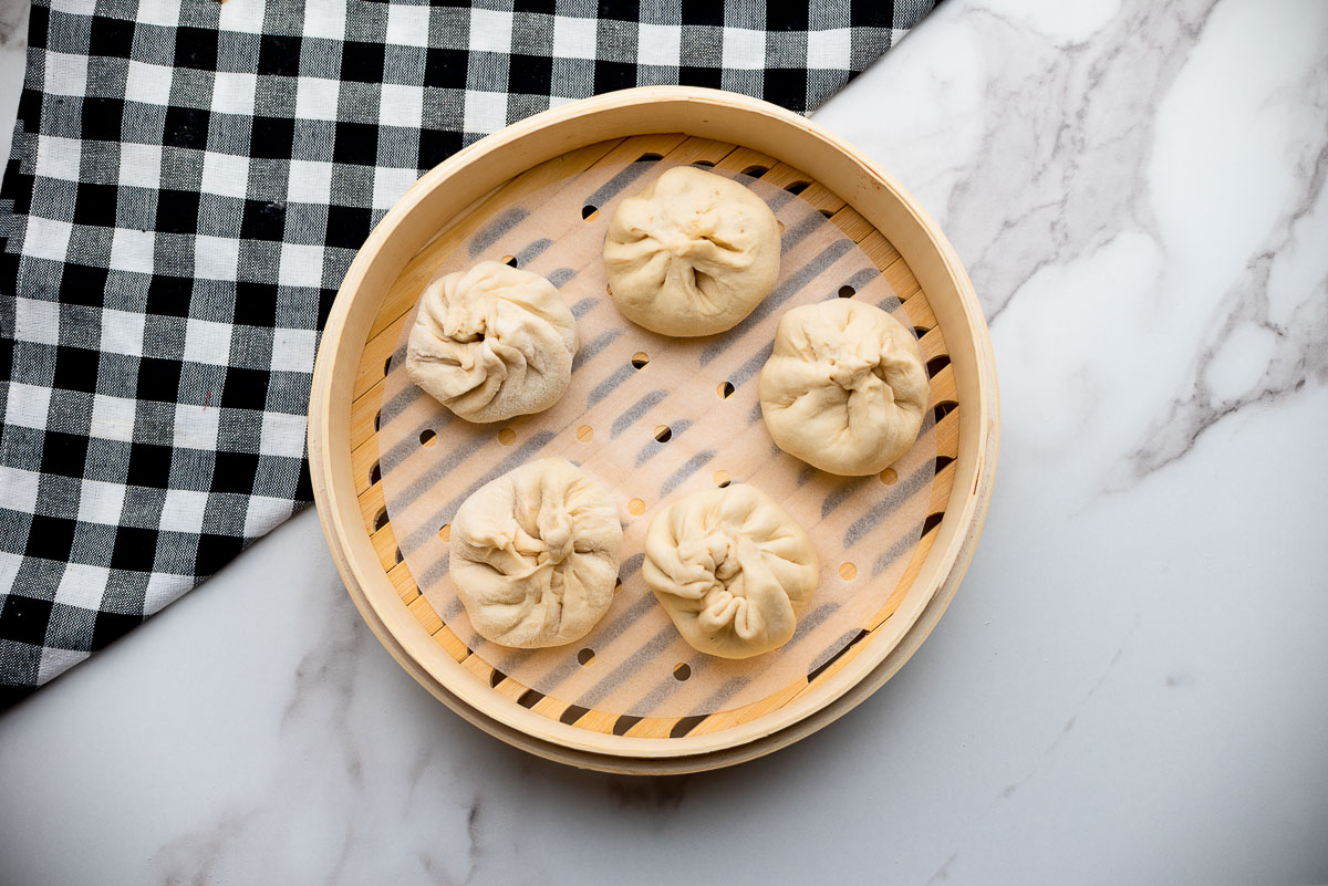 Five pork dumplings in a bamboo steamer ready to be cooked, placed on top of a marble surface with a black and white checkered napkin on the side.