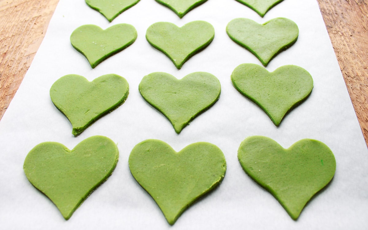 Green heart-shaped matcha sugar cookies on parchment paper ready to be baked, on top of a wooden board.