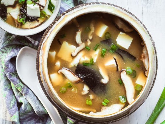 A bowl miso soup with tofu and shiitake mushrooms with sliced green onions and a white spoon on the side.