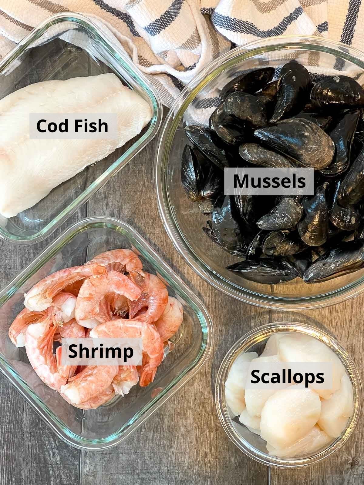 Fresh cod fish, mussels, shrimp, and scallops in separate glass bowls on top of a wooden plank board.