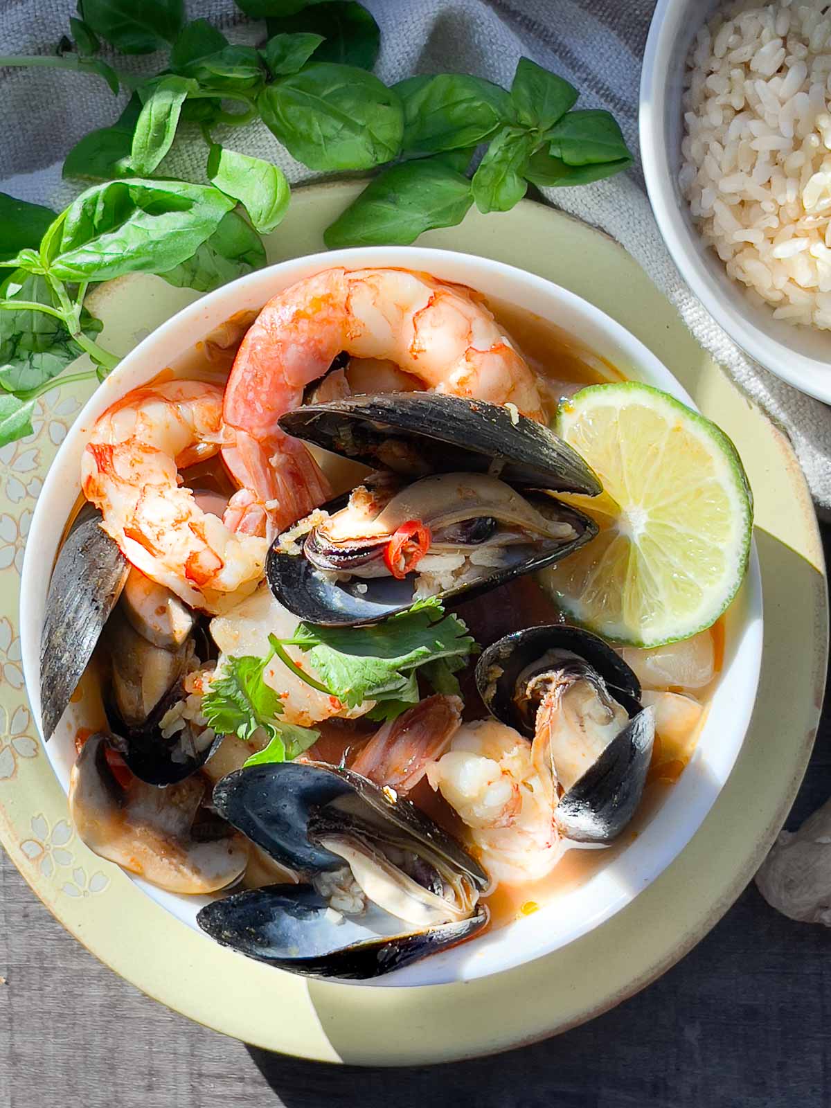 A white bowl of Thai seafood soup with shrimp, mussels and lime on top of a yellow plate with fresh herbs and a small bowl of rice on the side placed on top of a wooden board.