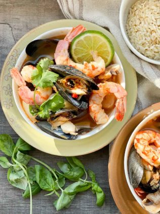 A white bowl of Thai seafood soup with shrimp, mussels and lime on top of a yellow plate with fresh herbs and a small bowl of rice on the side placed on top of a wooden board.