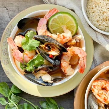 A white bowl of Thai seafood soup with shrimp, mussels and lime on top of a yellow plate with fresh herbs and a small bowl of rice on the side placed on top of a wooden board.