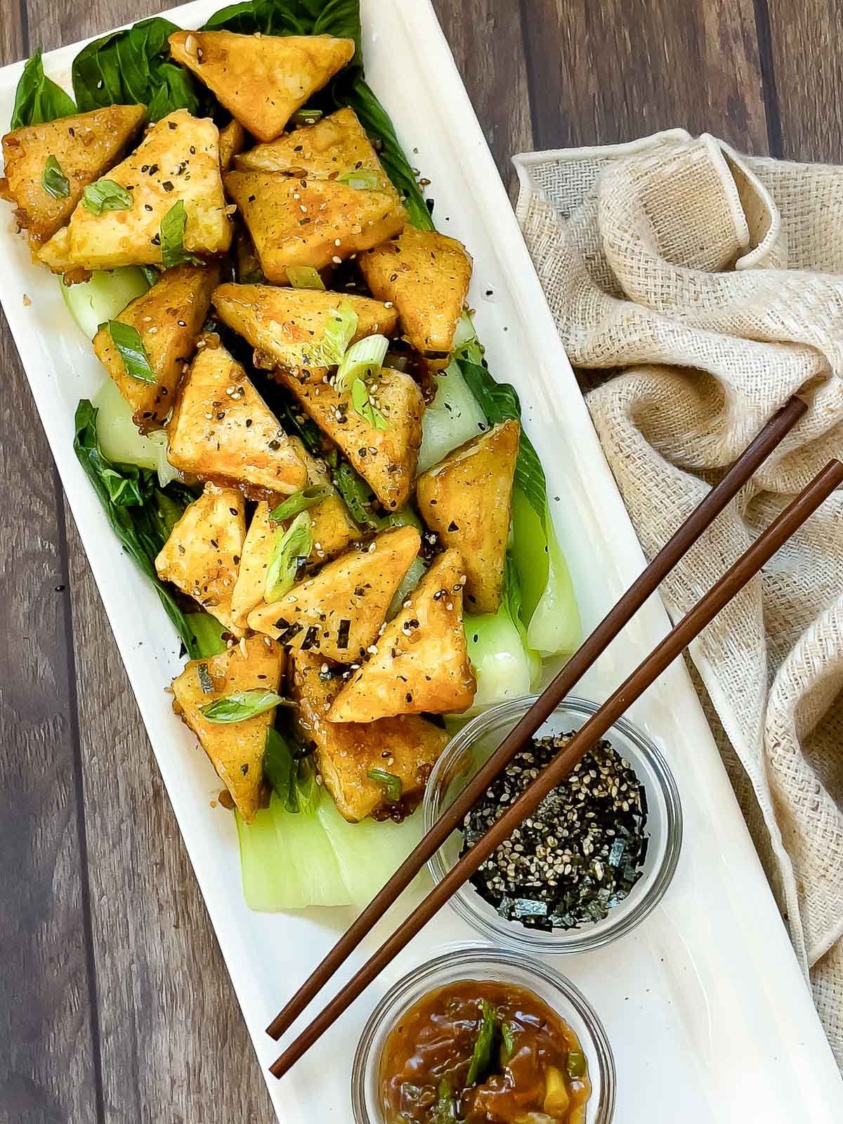 Golden-brown, crispy tofu steaks piled on top of cooked baby bok choy and sprinkled with seasonings plated on top of a white serving platter with small bowl of sauce and furikake seasoning on the side, and a pair of chopsticks on top and a napkin on the side.