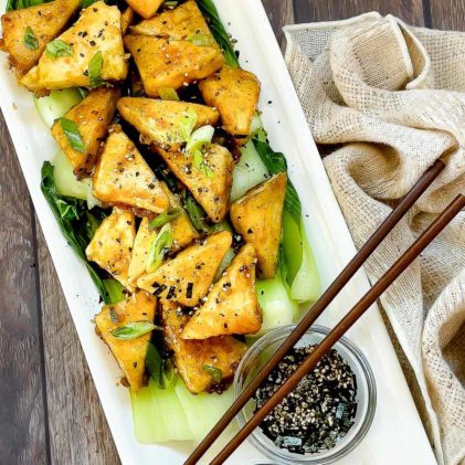 Golden-brown, crispy tofu steaks piled on top of cooked baby bok choy and sprinkled with seasonings plated on top of a white serving platter with small bowl of sauce and furikake seasoning on the side, and a pair of chopsticks on top.