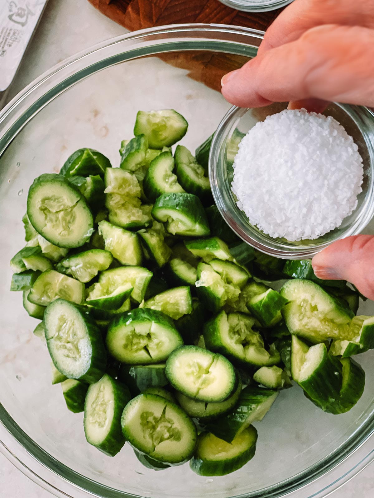Sliced cucumbers in a glass bowl with a small bowl of salt held above it.