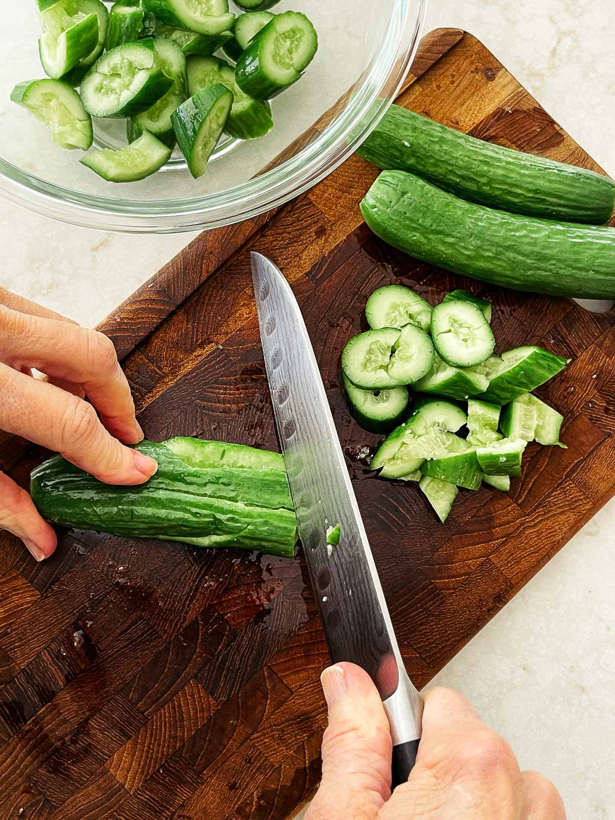 Persian cucumbers being sliced on top of a wooden cutting board with a glass bowl filled with sliced cucumbers on the side.