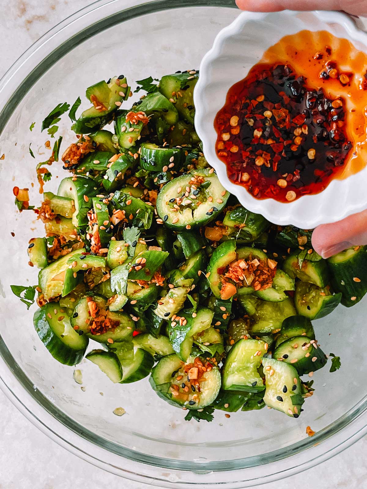 Smashed cucumber salad in a clear glass bowl with a white bowl of chili oil being poured on top.