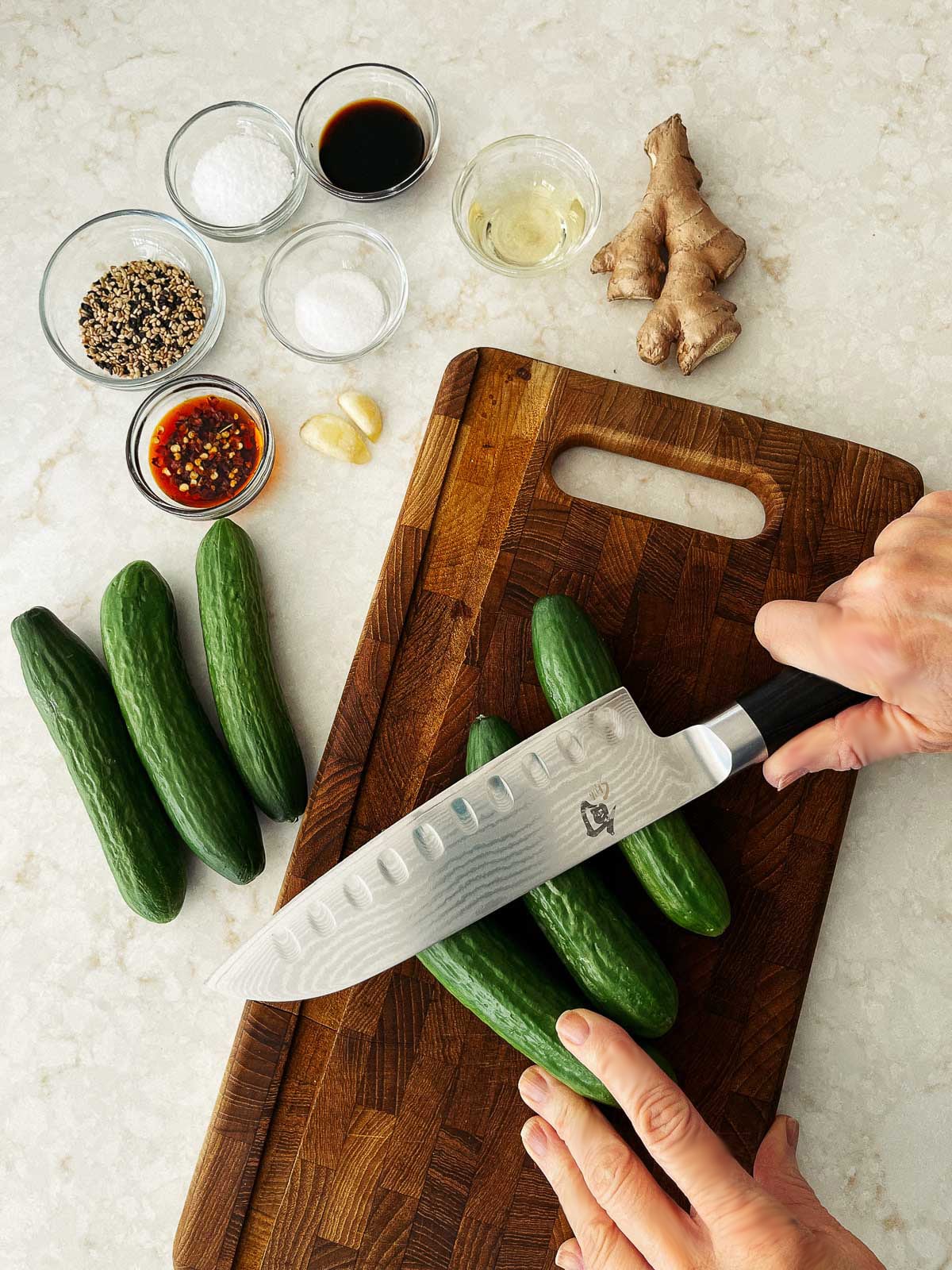 Persian cucumbers being pounded with a chef's knife on top of a wooden cutting board with more cucumbers and other ingredients on the side.
