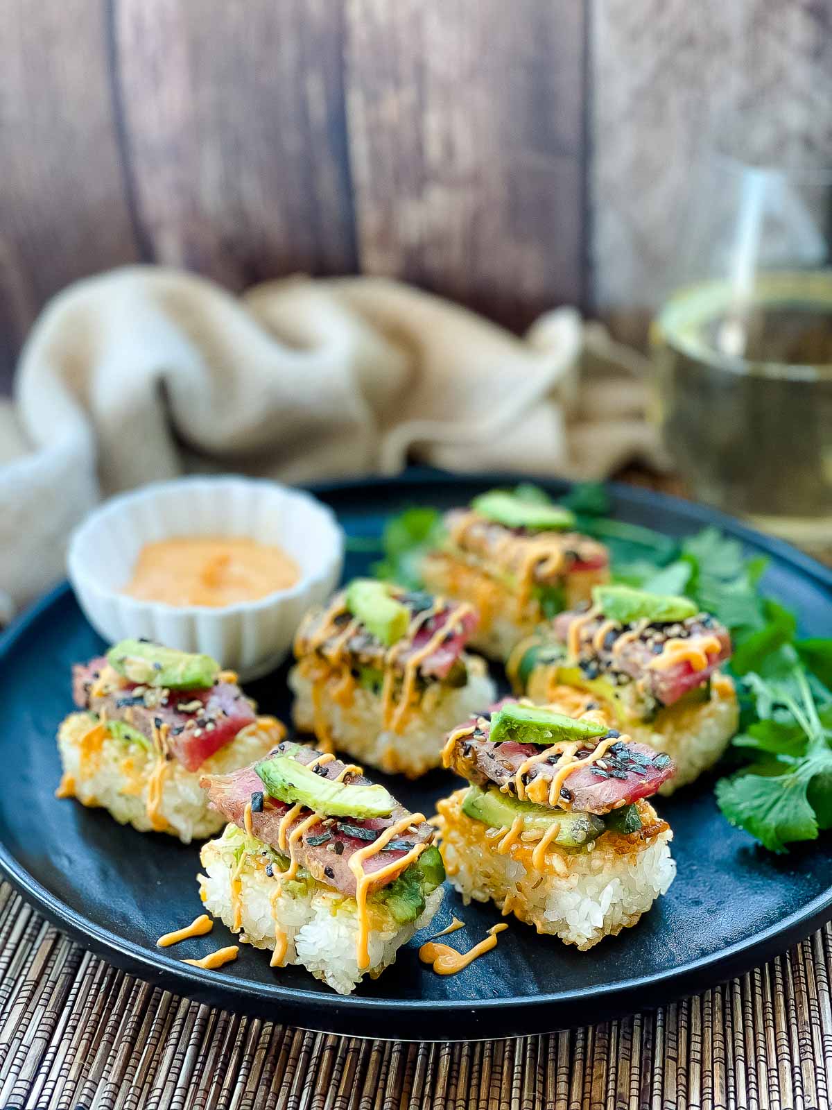 Spicy tuna crispy rice bites topped with avocado and tuna sushi drizzled with a sriracha aioli placed on top of a black plate with a small white bowl of aioli on the side and a napkin and glass of white wine in the background.