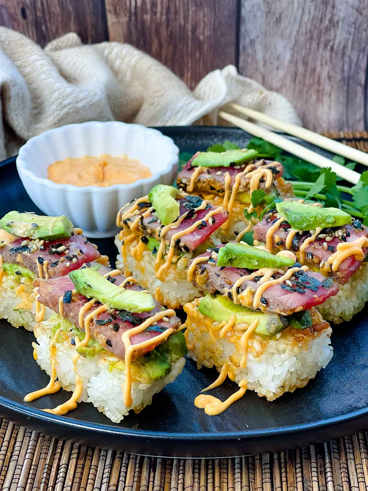 Spicy tuna crispy rice bites topped with avocado and tuna sushi drizzled with a sriracha aioli placed on top of a black plate with chopsticks and a small white bowl of aioli on the side