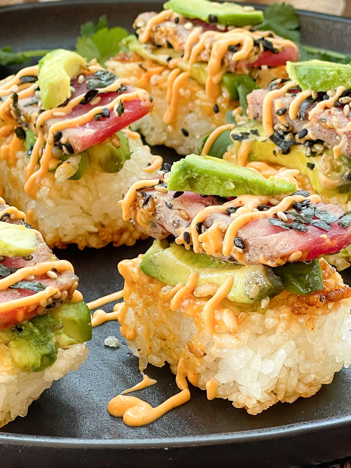 Spicy tuna crispy rice bites topped with avocado and tuna sushi drizzled with a sriracha aioli placed on top of a black plate.