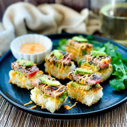 Spicy tuna crispy rice bites topped with avocado and tuna sushi drizzled with a sriracha aioli placed on top of a black plate with chopsticks and a small white bowl of aioli on the side