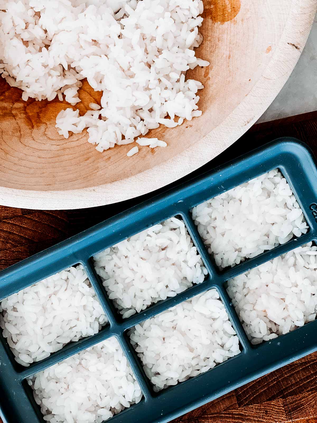 A black silicone ice cube tray filled with sushi rice and a wooden bowl of rice on the side.