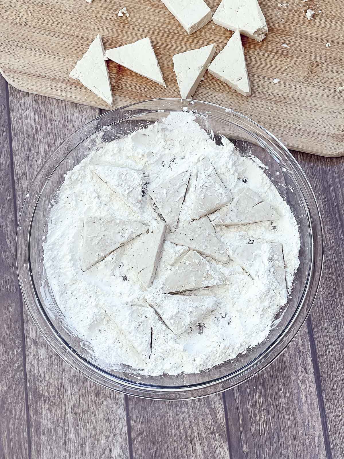 Tofu triangles being dredged in corn starch in a clear glass pie plate on top of a wooden board.